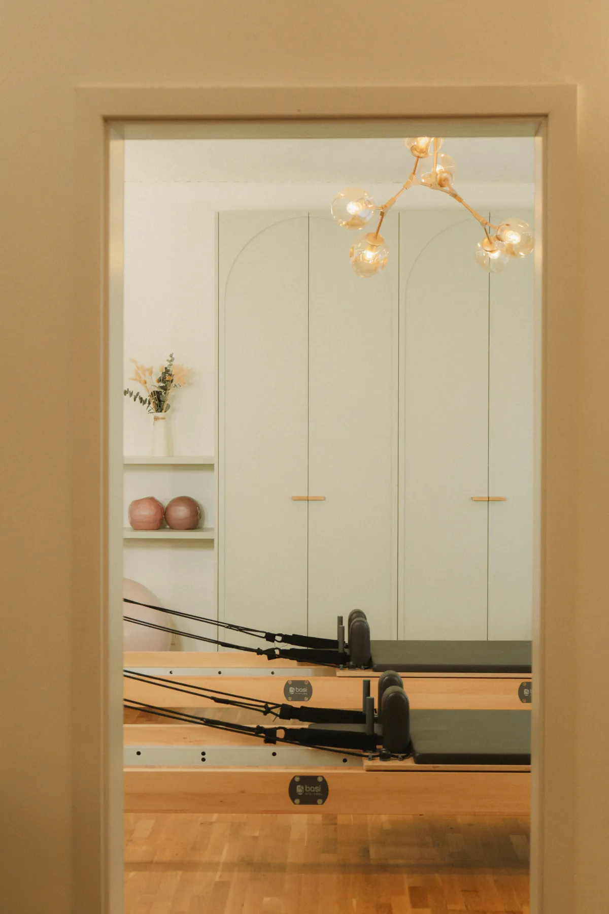 Pilates Studio Munich: reformers behind sage-green arched joinery and a brass globe chandelier