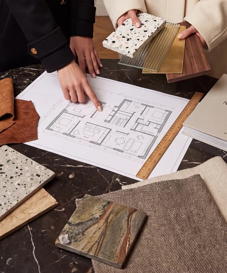 Two people over a floor plan with terrazzo, brass, timber and stone samples on a marble table